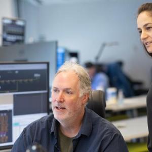 A person sat on a chair looking over a computer screen, while another person stands beside them