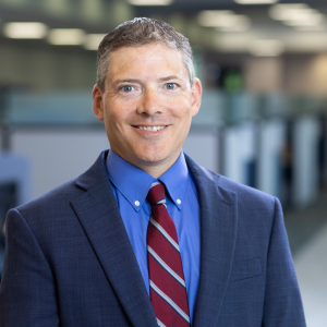 Man wearing a navy suit, blue shirt, and red striped tie, smiling in an office setting with cubicles in the background.