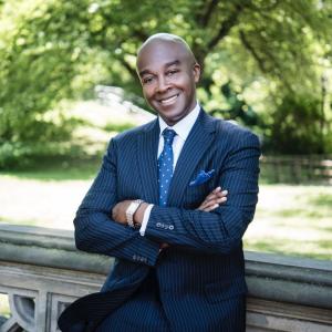 The color photograph shows Michael Carby, of Jamaican heritage leaning against a stone railing located outdoors. He’s wearing a blue pin stripped suit, with a blue polka dot tie, a blue handkerchief and a watch. His arms are crossed and he’s smiling at the camera. It’s a sunny day with green grass and green trees and bushes in the background.