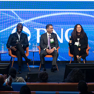 Five people seated on a stage with a huge digital display behind showing the PNC logo with blue background. All are smiling or laughing.