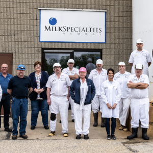 A small group of people posed outside a building. Some in all-white uniforms. "Milk Specialties" sign behind them. 
