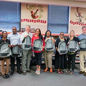 A line of people in a school setting, each holding a grey backpack.