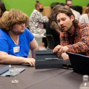 Two people looking at an open laptop in a room of others seated at different tables.