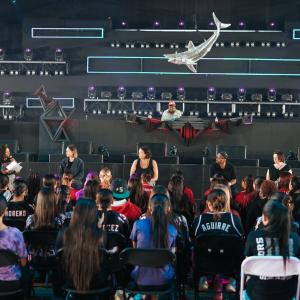A panel of speakers sits on stage facing a large seated audience inside a festival-style venue, with stage lighting rigs and a sculptural element hanging above.