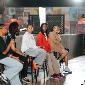 Six panelists sit on stools on a stage holding microphones, facing an audience off-camera, with venue artwork and lighting visible in the background.