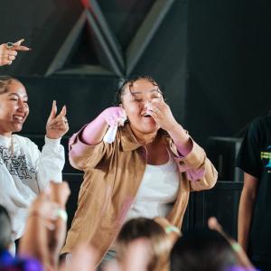 Three performers stand at the front of a stage engaging with a crowd; one holds a microphone while others gesture toward the audience, with hands raised in the foreground.