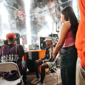 Several people sit and stand around audio and video equipment inside a reflective booth, looking toward a laptop screen displaying sound or video controls.