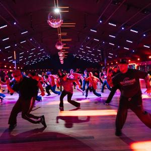 A large group of dancers moves across a wide dance floor under colorful lights and disco balls inside an indoor venue, captured mid-routine.