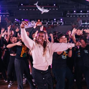 A person in a light jacket holds up a phone for a selfie while surrounded by a large group dancing with raised arms inside a concert-style venue with stage lighting overhead.