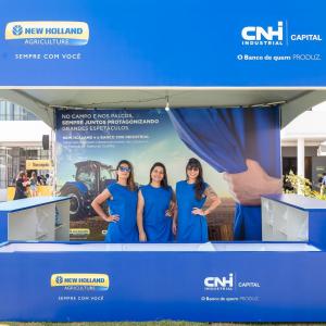 Three people in blue shirts at an outdoor covered booth with signs in blue for New Holland and CNH