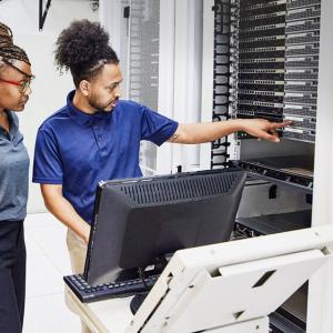 Two people in a room full of computer servers, a screen and keyboard on a small table in front of them.