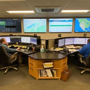 two people seated at desks with multiple monitors in front of them and mounted on the wall in front of them. Maps and data showing on the screens.