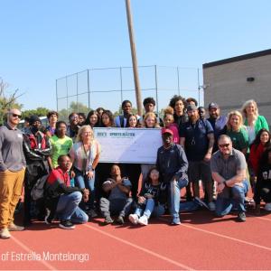 A group of people on a running track, some holding a large check. "Photo Courtesy of Estrella Montelongo" in the corner.