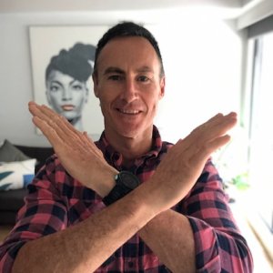 Man in a plaid shirt, standing in front of a picture of a woman, crossing his arms