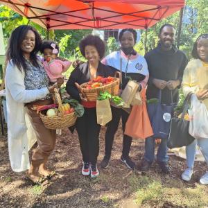 A group of people including a young child being held, each carry bags and baskets of fresh produce. Standing under a red pop-up awning outside.
