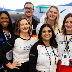 group of six people standing with planes in the background. One holds a small rainbow flag.