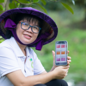 Nguyen Thi Kim Thoa sitting in an orchard showing a cellphone to the camera. She is wearing a large rimmed hat.