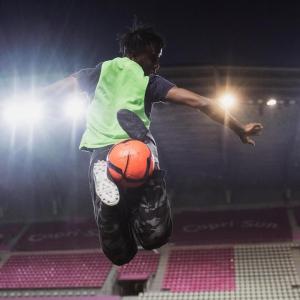 The back of a person doing a soccer trick move with the ball between their feet mid air. A lit stadium in front of them.