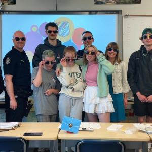 A group of adults and kids all wearing rainbow-rimmed glasses in a classroom setting. Some adults in police uniform.