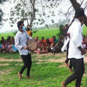 a large group of people outside, spectators sitting under trees while some performers are central, one playing a drum