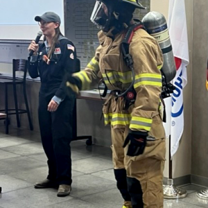 A person dressed in full fire-fighting PPE and a person next to them with a microphone.