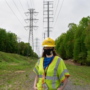 PSEG female employee standing under high tension power lines wearing protective gear, helmet and mask.