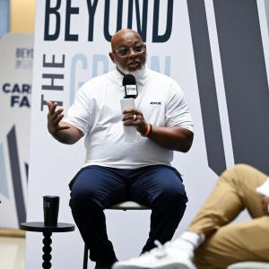 A person seated with a microphone talking. Another to the side. "PGA Works Beyond the Green" behind them.