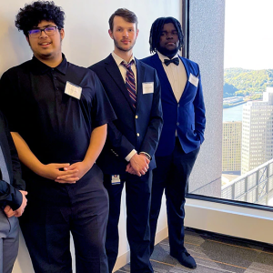 Five students in business style clothing standing in a hallway with a large window at the end overlooking a city landscape.