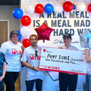 Group of people stood in front of a wall with red, white and blue balloons holding a Kentucky Fried Wishes funding cheque