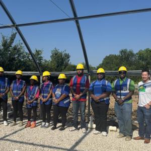 Group of people in greenhouse structure, wearing blue construction vests and yellow hard hats