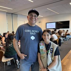 An adult and a youth participant stand together in a workshop space, with other attendees seated at tables behind them during a group program.