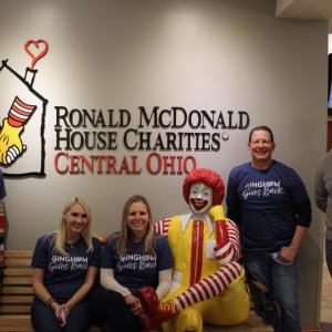 Five Bath & Body Works associates pose for a picture in front of a wall with a large logo of the Ronald McDonald House Charities of Central Ohio on it. Three associates are standing and two are sitting on a bench that includes a statue of Ronald McDonald with his leg crossed and showing a big smile.