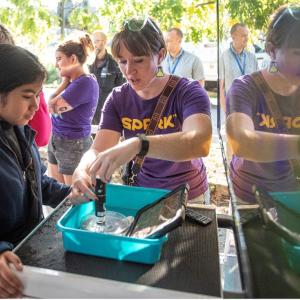 A woman in a purple SPARK MilliporeSigma t-shirt demonstrates an experiment to a student on the Curiosity Cube. 