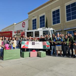 A mix of team members from Qurate Retail Group are standing outside a QVC facility in front of a firetruck along with fire fighters. A few people are holding up a banner which reads "QVC Do What You Love, Love What You Do". They are also surrounded by wrapped gift boxes and holding children's toys as part of a holiday toy drive event.  