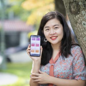 Quynh holding up a phone, leaning on a tree.