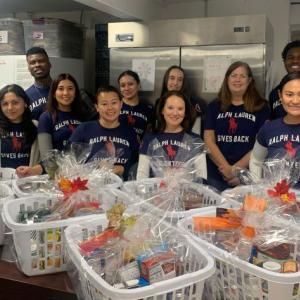 A group of people standing in a kitchen, large baskets of food items in front of them. All wearing "Ralph Lauren Volunteer" t-shirts.
