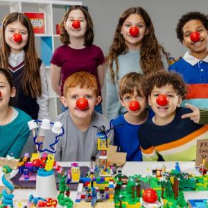 Children wearing red noses behind a table with lego models on 
