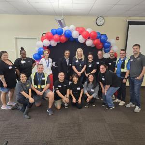 A group of men and women posing for a photo in front of a black backdrop with an arch of red, white, and blue balloons overhead.