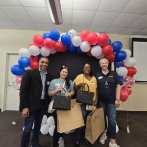 A man and three woman posing for a photo in front of a black backdrop with an arch of red, white, and blue balloons overhead.