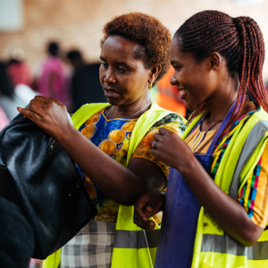 A person inspecting a handbag as another smiles, looking on.
