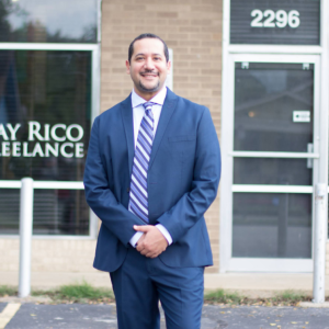 Ray Rico standing outside their business. Wearing a blue suit and striped tie.