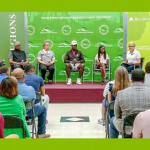 A panel of Black leaders, athletes, and students on a raised platform in front of a green backdrop