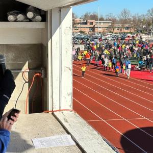 Robert McNamee in a booth with a microphone, looking out to a race track filled with participants.