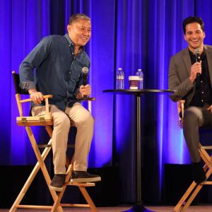 Two people on a stage in directors chairs, holding microphones.