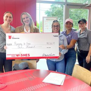 Group of people stood in front of a red wall holding a Kentucky Fried Wishes funding cheque