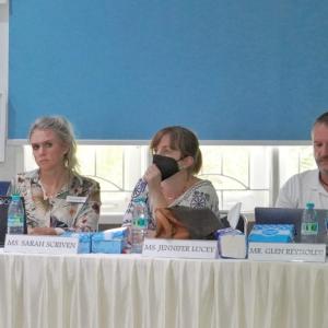 Three people at a desk with name placards, two with laptops open.