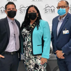 Three people wearing protective masks in front of a wall with SIM logos