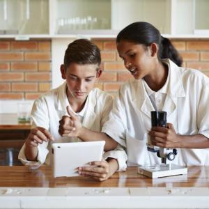 Two young students in a lab looking at a tablet device while another holds a microscope
