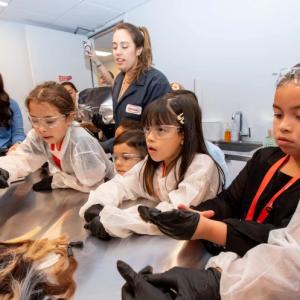 Children and adults in protective lab gear surround a table.