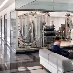 People walking in a hallway and seated in a reception area. A person in protective lab gear behind a glass-walled room.